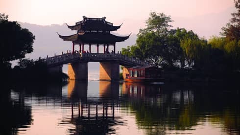 A boat floats towards a stone bridge with a pagoda cover cast in orange light from the setting sun.