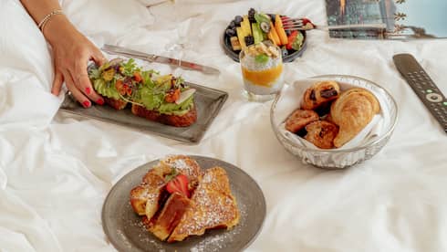A few small plates of food items like pastries and French toast are arranged on a bed, next to a magazine and remote control. A person's hand reaches for one of the food items.