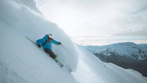 Skier in blue jacket, helmet skis down slope, snow spraying behind him