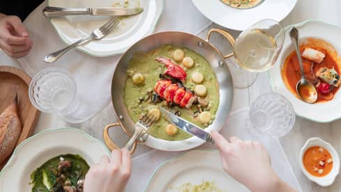 Gourmet meal featuring lobster, pasta, and green sauce served in a pan, with various dishes and a glass of white wine on a table, as someone cuts into the food