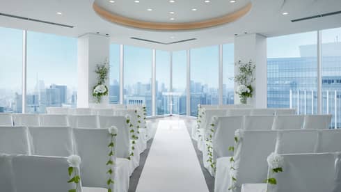 An indoor wedding venue with rows of white chairs and flowers, with a view of the city skyline from window.