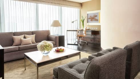 Living area with grey sofas, coffee table with flowers and fruit, work desk with a chair and floor-to-ceiling windows with curtains