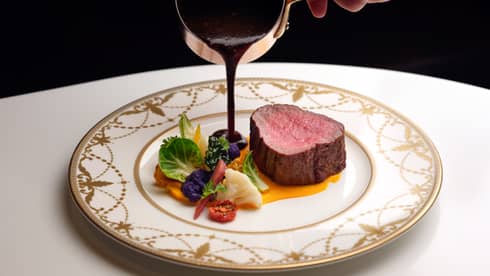 Thick slice of beef fillet on a white and gold plate, as person's hand pours over a gravy sauce