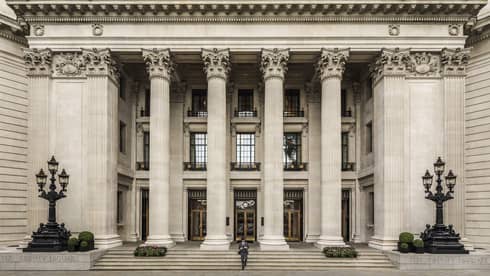 Man in suit and black hat walks down steps under tall white pillars of building