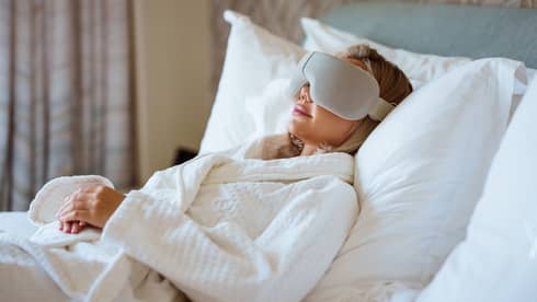 Person wearing white spa robe lays back on a bed with large white pillows wearing a high-tech eye mask
