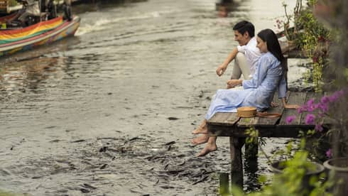 Couple sits on the dock of the Chao Phraya River, hungry fish swarming beneath their dangling feet