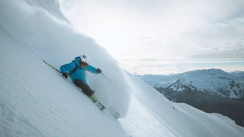 Skier in blue jacket, helmet skis down slope, snow spraying behind him