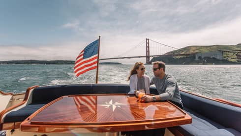 A man and woman on the back of a boat with a metal bridge in the distance behind them and an American flag on the boat.