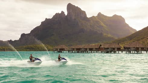 Two jet skis ride along lagoon past row of overwater bungalows, Bora Bora mountain
