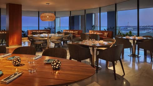Indoor restaurant dimly lit, wooden tables and brown chairs, wall of windows