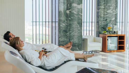 Two guests dressed in white bathrobes lounging on recliners in a modern spa, with two mugs of tea on a table in between them