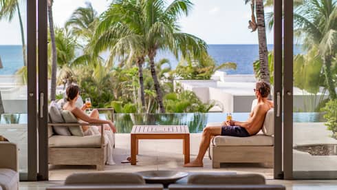 Two people relaxing with drinks on a terrace overlooking palm trees and the ocean.