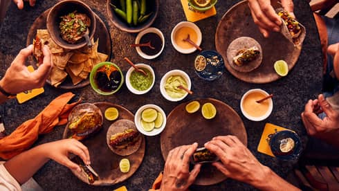 An overhead view of five pairs of hands holding tacos next to bowls of chips, salsa, peppers, limes and sauces.