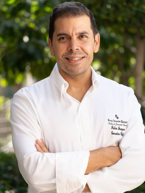 A chef in a Four Seasons uniform with his arms crossed in front of a canopy of foliage.