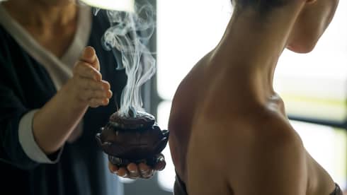Smoke rises from decorative wood bowl in hotel staff's hands, against woman's bare back