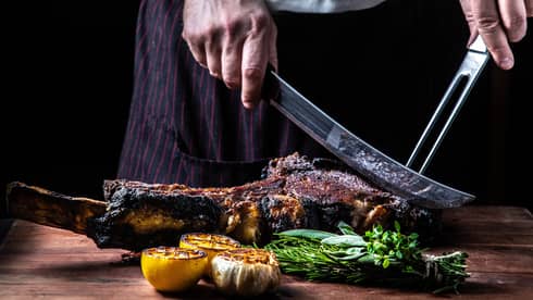 Close-up of chef slicing large rack of grilled meat, vegetables with butcher knives