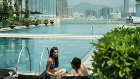 Two people are by the pool edge, and the city skyline is behind them. The pool area has greenery, lounge chairs and plants and there is a clear sky overhead.