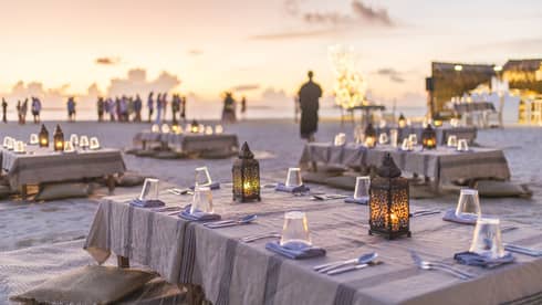 Dining tables with cloths and candles on a beach, a crowd of people in the distance.