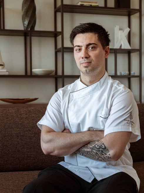Pizza specialist DeLeo wearing white chef's coat sits on couch with arms crossed