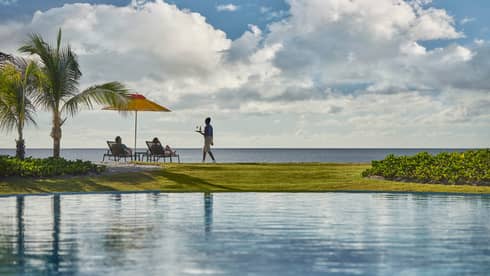 View across pool to green lawn, waitstaff carrying tray to couple in patio chairs overlooking ocean