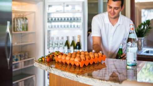 Smiling man holds green bottle in front of open fridge, fresh eggs on counter