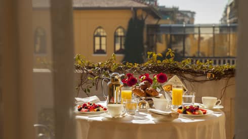 Breakfast pastries, juice, coffee and red flowers on small private dining table on sunny balcony