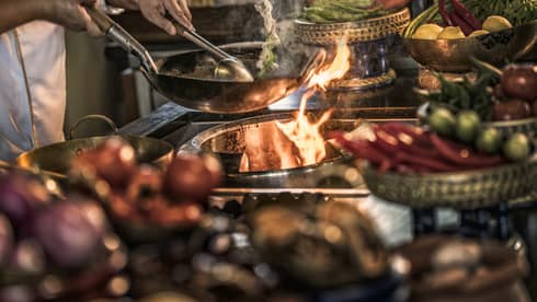 Close-up of chef stirring meals in copper pans over stove, flames 