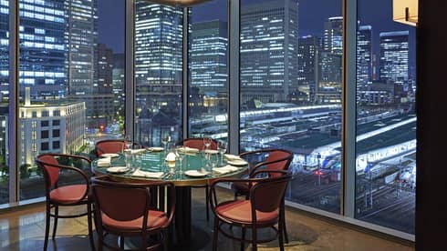 Luxury boutique hotel dining setup with a round table elegantly set for guests, offering a panoramic city view at night through tall glass windows