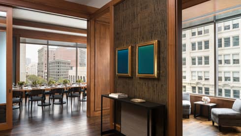 A waiting area in a restaurant with grey chairs and a small table, a private dining area with large windows can be seen through a hall.