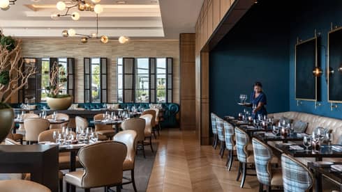 The interior of cinderhouse restaurant at four seasons saint louis is decorated with a navy accent wall, light wooden floor with a chevron style, black tables and camel colored leather chairs
