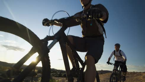 Close-up of cyclist in gear and helmet looking up from bike, second cyclist nearby as sun shines from behind