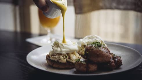 Hollandaise sauce being poured onto eggs Benedict with lump crab meat, side of crispy roasted potatoes 