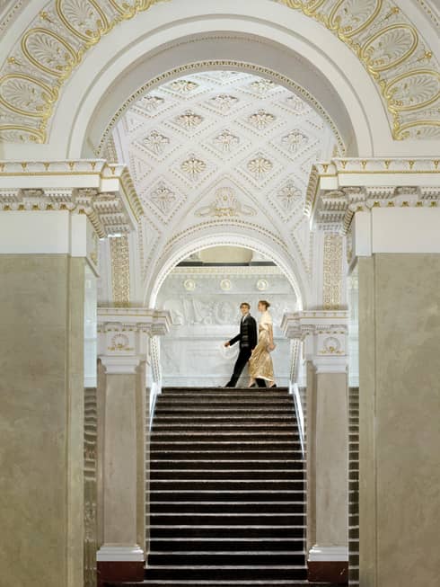 Woman in gold evening gown with fur jacket and man in suit walk at top of marble staircase