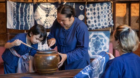 A woman teaching a child how to dye fabric using indigo, with tie-dyed cloths hanging in the background in a traditional workshop.