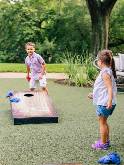 A small boy and girl playing corn hole near a large tree outside.