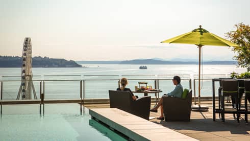 Couple sits poolside at dining table on sunny patio overlooking Elliott Bay and The Seattle Great Wheel