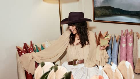 A person wearing a black wide-brimmed hat, beige blouse and white skirt holds a drink while examining a display of boots.