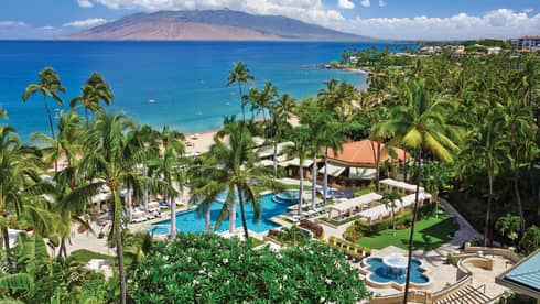 Aerial view of resort pool, patios and palm trees on ocean, mountain in background