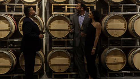 A couple in evening wear hold glasses of red wine and listen to a sommelier while looking at a rack of wine barrels.