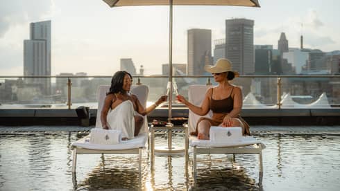 Two guests have a toast with drinks in stemmed glasses, while they sit on lounge chairs under a sun umbrella over a rooftop pool, with city skyline views in the background.