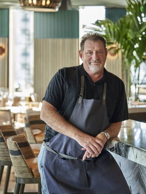 A chef in a blue apron leaning on a counter.