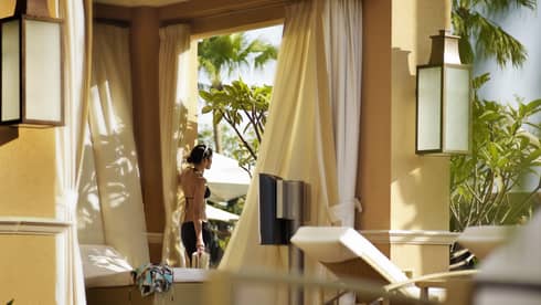 Woman wearing black bikini stands by padded patio bed under white cabana on sunny deck