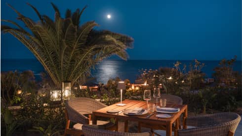 Dining table set for four on an outdoor patio at night with palm trees and the ocean in the distance