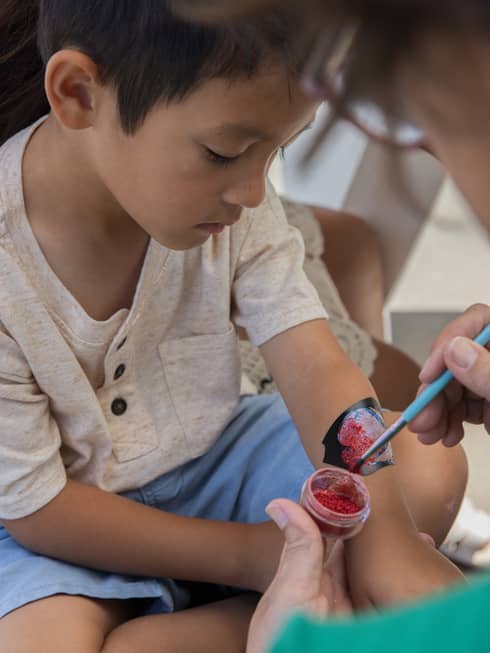 Child wearing beige shirt and light blue shorts sits getting their arm painted