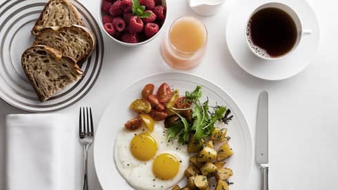 Breakfast plate with sunny-side-up eggs, cherry tomatoes, greens, and roasted potatoes, alongside toast, raspberries, grapefruit juice and coffee.