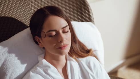Guest with long, light brown hair sits with their eyes closed on a spa lounge chair wearing a white robe