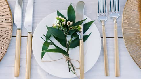 A bohemian table setting of gold chargers, modern long gold and silver flatware, white porcelain plates and a sprig of greenery
