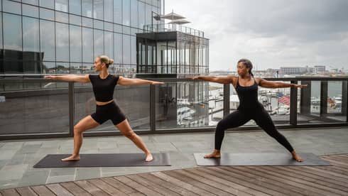 Two people stand in a yoga pose on a rooftop deck, with city views in the background.