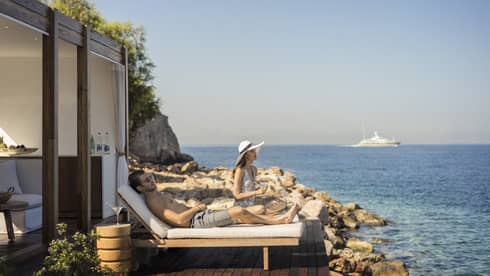 A couple sunbathes on the deck outside a private furnished cabana, only the rocky shore, the sea and a distant boat in view.