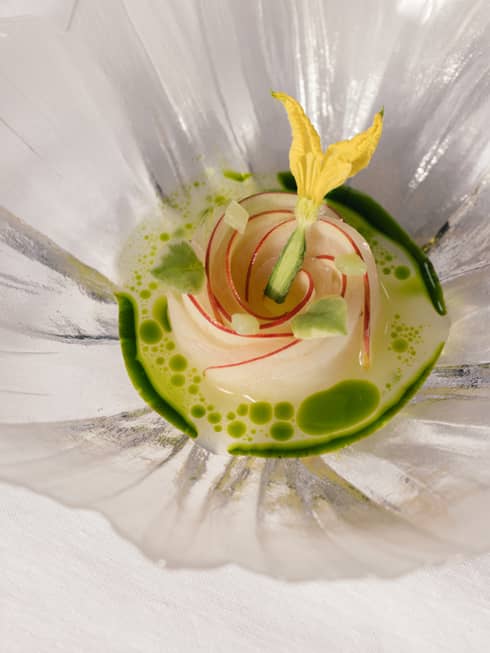 Elegant dish arranged as a rose, topped with a yellow edible flower, in a green herb-infused broth